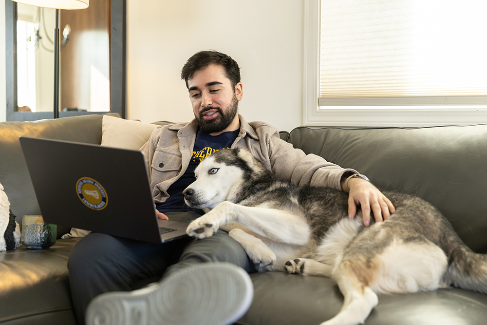A person sitting on a couch with a laptop computer, while a dog is lying next to them. The person has a beard and is wearing a gray jacket. The dog appears to be a medium-sized husky or similar breed. They appear to be relaxing and working on the laptop together.